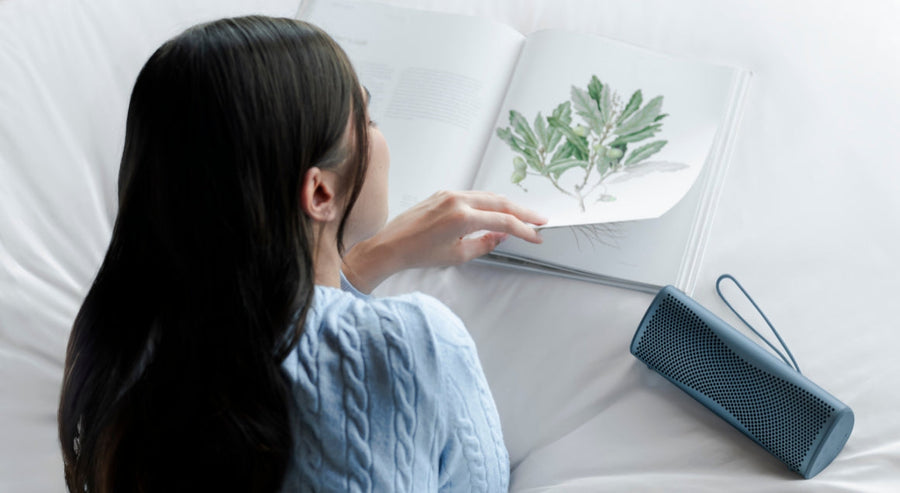 Person reading an illustrated book on a bed with a portable speaker placed nearby.