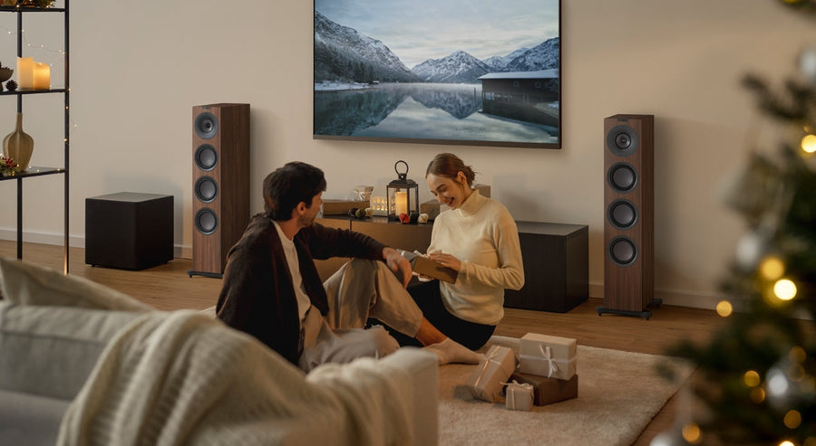  Couple sitting on the floor exchanging Christmas gifts in a cosy living room with wooden floorstanding speakers, a large TV, and soft festive lighting.