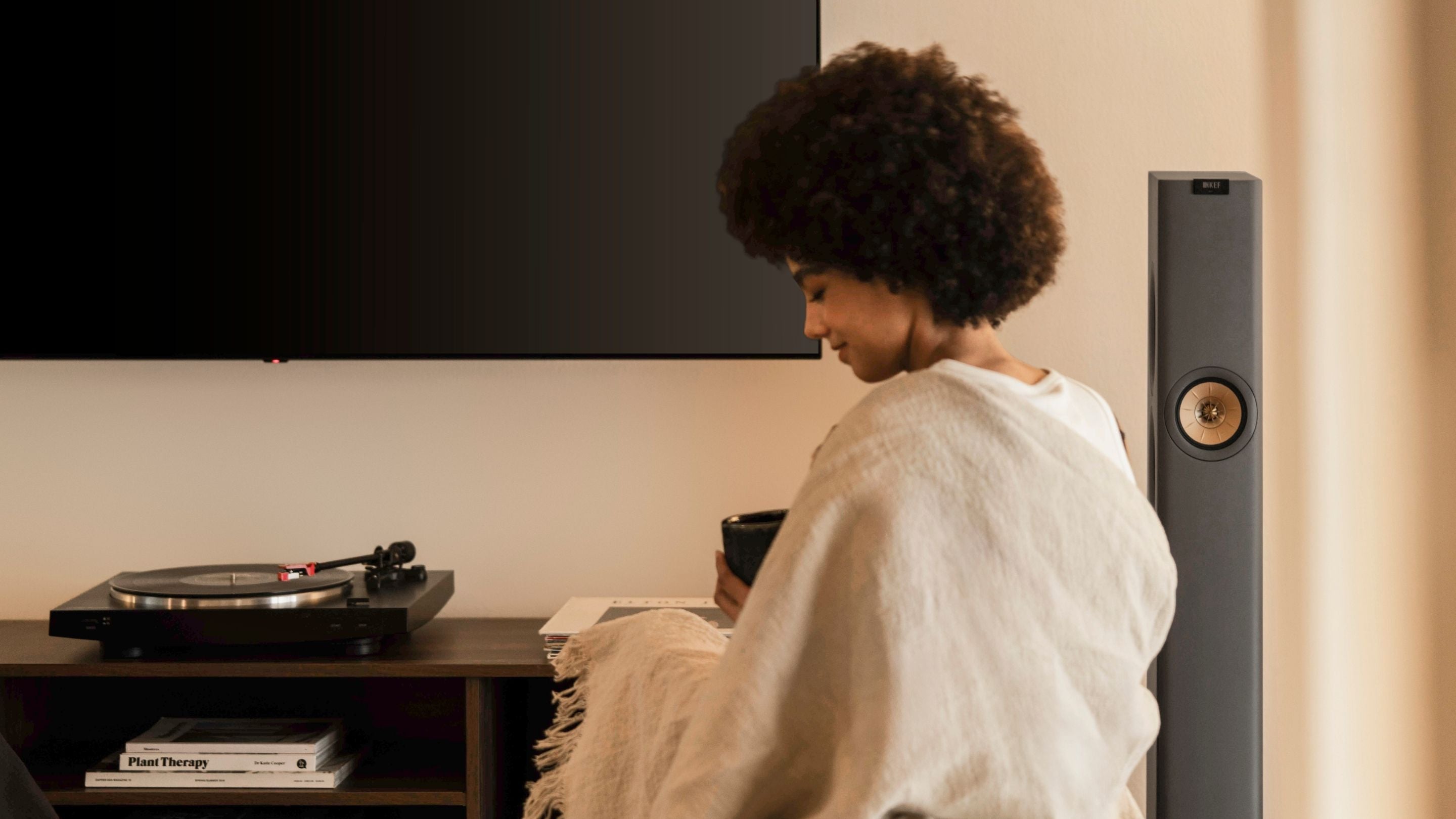 Person relaxing with a cup of coffee in a cosy living room, sitting near a KEF floor-standing speaker and a record player.