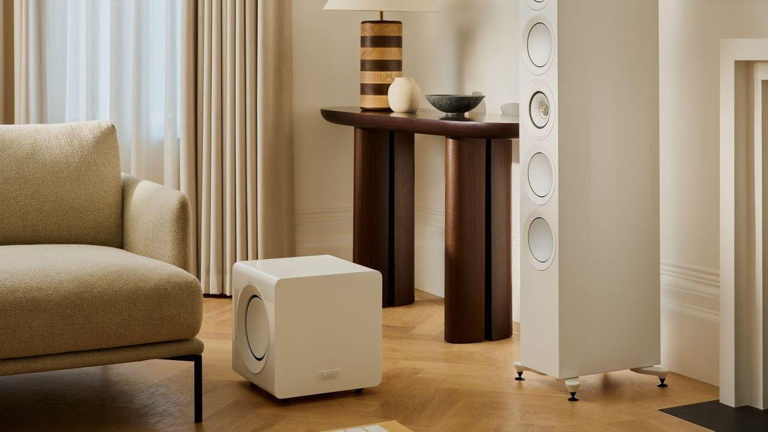 A living room setup with a comfortable chair, a small table, and a KEF KC92 White subwoofer next to a R11 Meta HiFi speaker