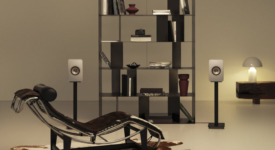 Modern minimalist living room with two white bookshelf speakers on black stands, a black open shelving unit with books and decorative items, a designer lounge chair, and a soft ambient table lamp.